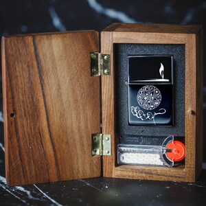 May include: A wooden box with a hinged lid, open to reveal a black lighter with a white design of a hand holding a circle with symbols. The lighter is in a black foam insert. The box has a brass hinge and a red knob.