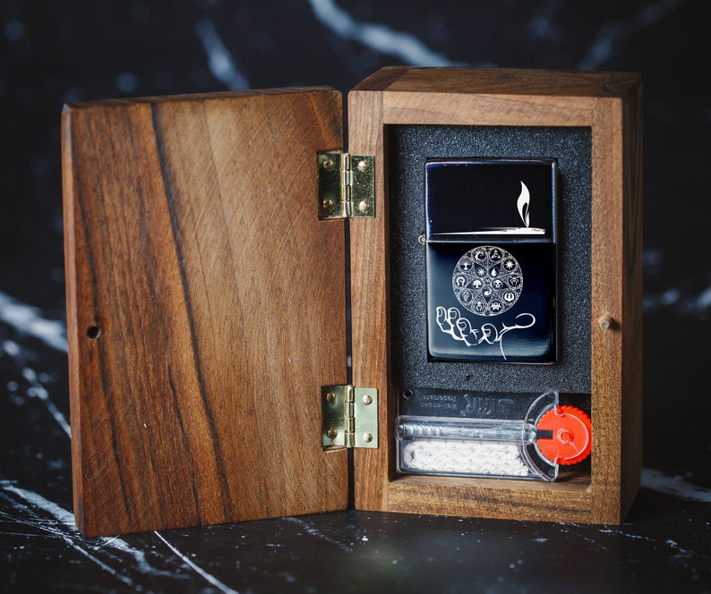 May include: A wooden box with a hinged lid, open to reveal a black lighter with a white design of a hand holding a circle with symbols. The lighter is in a black foam insert. The box has a brass hinge and a red knob.