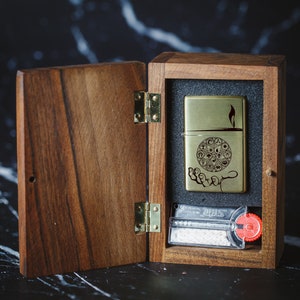 May include: A wooden box with a hinged lid, open to reveal a gold lighter with an engraved design and a small pack of flint. The lighter has a flame design and the text "Lighter".