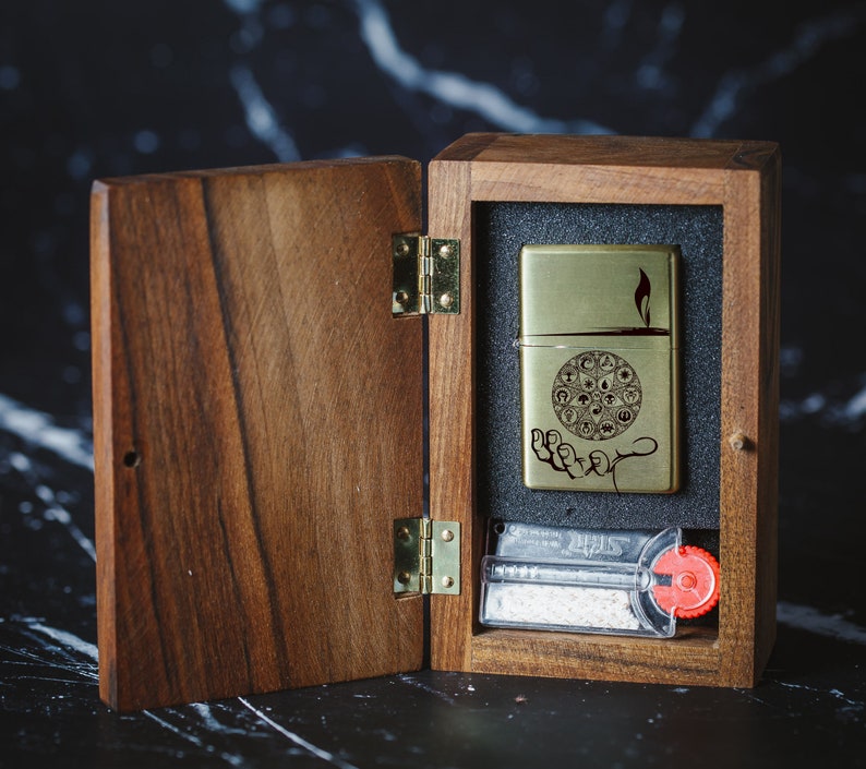 May include: A wooden box with a hinged lid, open to reveal a gold lighter with an engraved design and a small pack of flint. The lighter has a flame design and the text "Lighter".