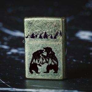 Puede incluir: Un encendedor de estilo vintage con un acabado verde met&aacute;lico desgastado. El encendedor presenta un dise&ntilde;o grabado de dos osos enfrentados en un entorno de bosque con estrellas y una luna en el fondo.