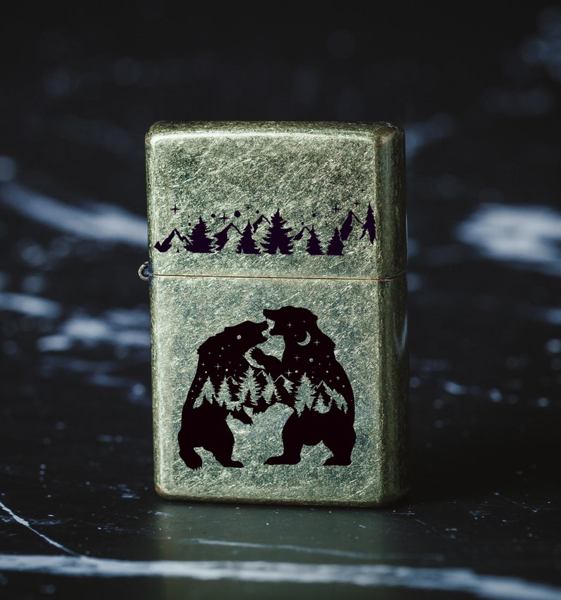 Puede incluir: Un encendedor de estilo vintage con un acabado verde met&aacute;lico desgastado. El encendedor presenta un dise&ntilde;o grabado de dos osos enfrentados en un entorno de bosque con estrellas y una luna en el fondo.