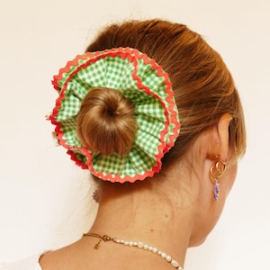 Puede incluir: Una scrunchie a cuadros verdes y blancos con un borde festoneado rojo se usa en un peinado de moño. La scrunchie es el punto focal, agregando un toque de color y patrón. El accesorio para el cabello es una adición lúdica.