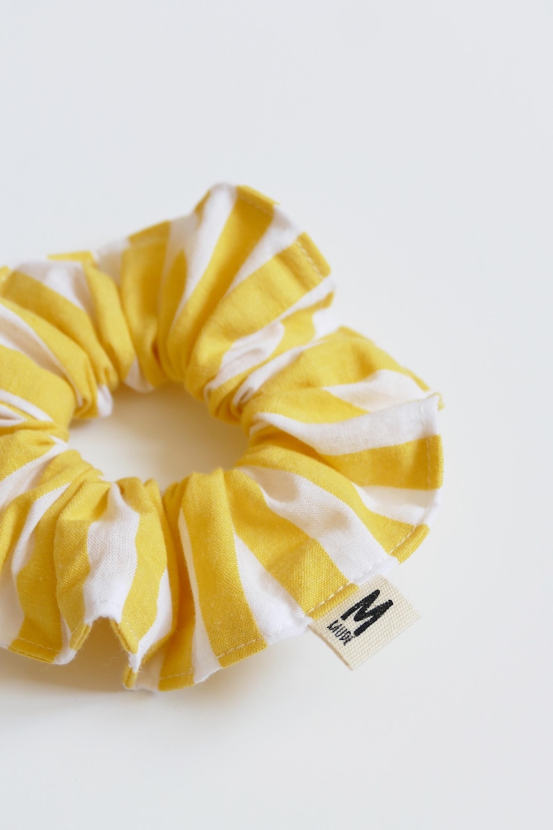 May include: A yellow and white striped scrunchie. The fabric is gathered and sewn into a circular shape. A small tag with the letter "M" and the word "LAUDE" is attached.