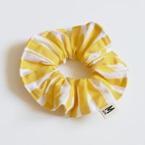 May include: A yellow and white striped scrunchie. The fabric is gathered around an elastic band, creating a ruffled effect. A small tag with the letter "M" is attached to the scrunchie.
