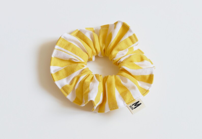 May include: A yellow and white striped scrunchie. The fabric is gathered around an elastic band, creating a ruffled effect. A small tag with the letter "M" is attached to the scrunchie.