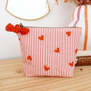 May include: A red and white striped cosmetic bag with red heart accents. The bag has a red bow attached to the zipper pull. The bag is on a wooden surface.