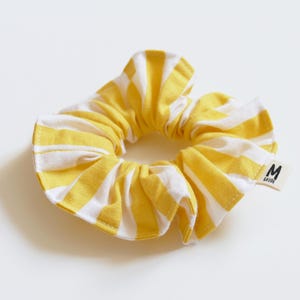 May include: Yellow and white striped scrunchie. The fabric is gathered around an elastic band, creating a ruffled effect. A small white tag with black text is attached to the scrunchie.