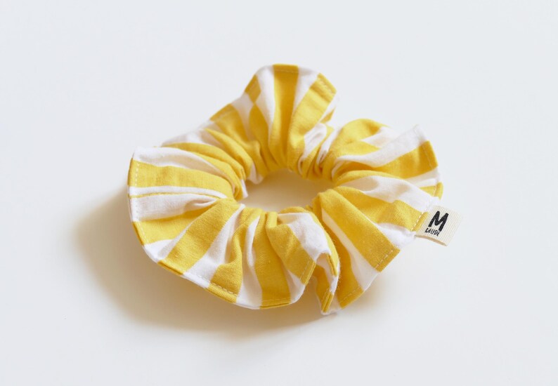 May include: Yellow and white striped scrunchie. The fabric is gathered around an elastic band, creating a ruffled effect. A small white tag with black text is attached to the scrunchie.