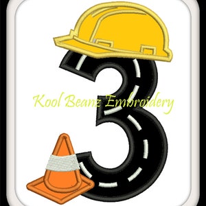 May include: A black number three shaped like a road with white lines. A yellow construction hat sits on top of the number. A small orange traffic cone is to the left of the number. The text "Kool Beanz Embroidery" is below the number.