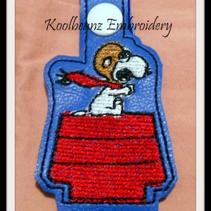 Puede incluir: Un llavero de cuero azul con un botón blanco y una imagen bordada de Snoopy con su bufanda roja de pie en su caseta. El texto "Koolbeanz Embroidery" está bordado en el llavero.
