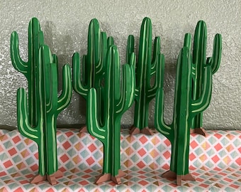 May include: A set of six green wooden cactus figurines. The cacti are painted in a bright green color and have a realistic shape. They are arranged in a row on a pink and white patterned surface.