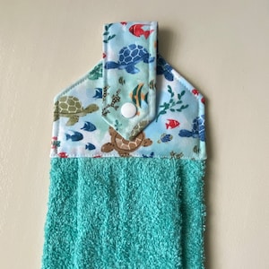 May include: A teal kitchen towel with a sea-themed print featuring turtles, fish, and coral. The towel has a loop for hanging and a white button.