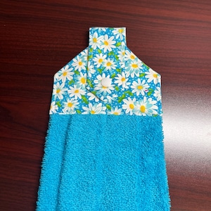 May include: A hanging kitchen towel with a blue terrycloth bottom and a daisy print top. The top features a blue background with white daisies and yellow centers. A white button secures the top flap.
