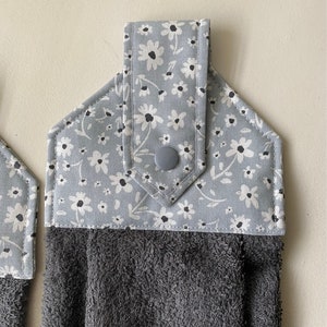 May include: Two gray kitchen towels with a floral print and a gray button on the top. The towels are hanging on a white wall.