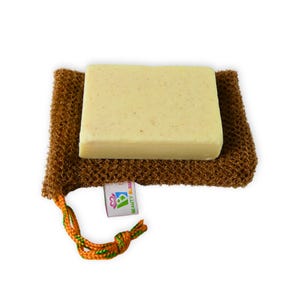May include: A rectangular bar of light-colored soap rests on a brown, textured washcloth. The washcloth has a drawstring closure with orange and green accents. A small tag with the text "BEAUTY BLISS" is attached to the washcloth.