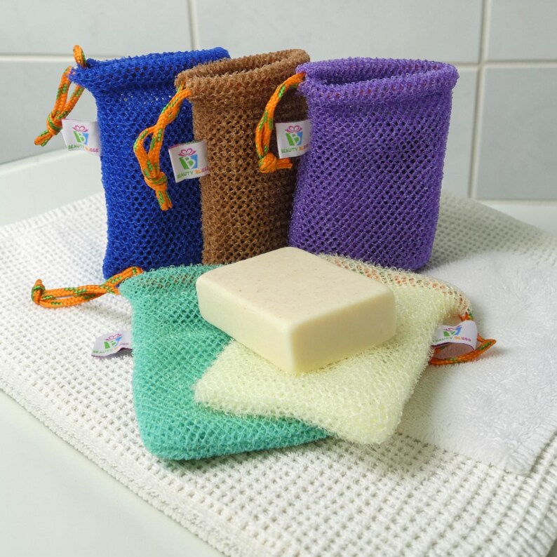 May include: A collection of colorful mesh soap bags in blue, brown, and purple, with a green and yellow bag holding a bar of soap. Each bag has a drawstring closure with orange and green cord. The bags are on a white waffle-weave towel.