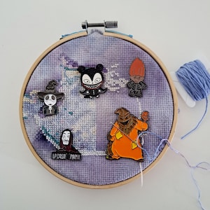 Könnte beinhalten: Ein lila-weißer bestickter Reifen mit verschiedenen Charakter-Pins. Die Pins zeigen ein schwarzes und weißes Skelett, eine schwarze und weiße Kreatur mit einer Krone, eine schwarze und weiße Kreatur mit einem roten Herzen, eine schwarze und weiße Kreatur mit einem roten Hut und eine schwarze und weiße Kreatur mit einem roten und orangefarbenen Outfit.