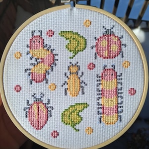 May include: A cross-stitch embroidery hoop featuring a design of colorful insects and leaves. The insects are in shades of pink, yellow, and orange, with green leaves and small orange dots. The embroidery is set in a wooden hoop.