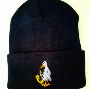 Puede incluir: Un gorro negro con dobladillo. El gorro presenta un diseño bordado de una gaviota blanca tocando un saxofón dorado. La gaviota tiene patas amarillas y un pico amarillo. El gorro está hecho de punto.