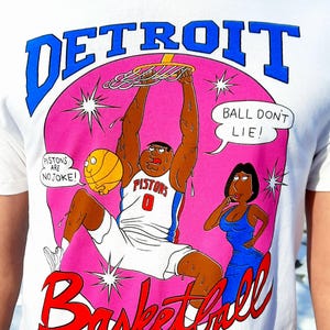 May include: White t-shirt with a Detroit Basketball graphic. The design features a cartoon basketball player dunking, a woman in a blue dress, and text that reads "Detroit Basketball" and "Ball Don't Lie!"