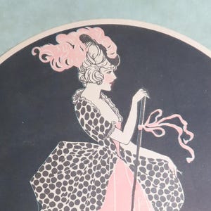 May include: Round black decorative plate featuring a vintage illustration of a woman in a pink and black polka-dot dress, holding a cane. The woman has a pink feathered hat and is set against a black background.