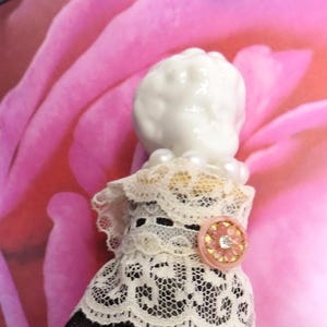 May include: A vintage-style doll with a white porcelain head and a delicate lace dress. The dress features a black mesh skirt with polka dots, a cream-colored lace bodice, and a pink button with a floral design. A string of pearls adorns the neck.