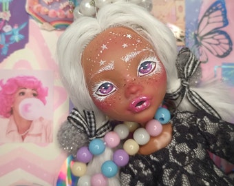 Peut inclure: Une poupée aux cheveux blancs et à la peau brune portant une robe en dentelle noire et une couronne de perles et de bonbons. La poupée a des ombres à paupières violettes et des lèvres roses. La poupée porte un collier de perles colorées.