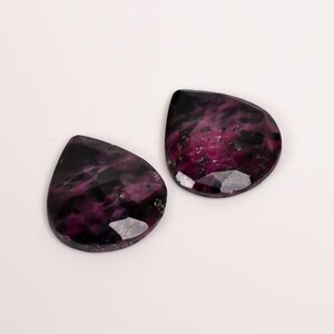 Könnte beinhalten: Zwei facettierte, tropfenförmige Edelsteine. Die Steine haben einen tiefvioletten Farbton mit schwarzen und grauen Marmorierungen. Die Edelsteine ​​sind vor einem weißen Hintergrund platziert und zeigen ihre polierten Oberflächen und einzigartigen Muster.