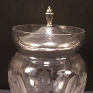 May include: A clear glass jar with a silver lid and etched floral design. The jar has a round body and a wide mouth.
