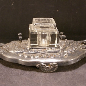 May include: A silver inkwell with an ornate design and a glass inkwell insert. The inkwell has two handles and sits on a base with a decorative design.