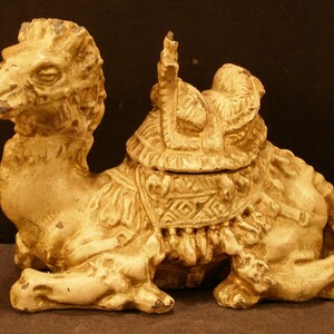 May include: A white ceramic camel figurine with a detailed saddle and ornate decorations. The camel is sitting with its head turned to the side and its legs tucked underneath its body.