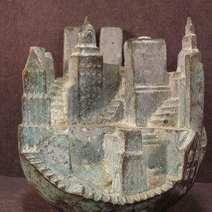 May include: A bronze-colored sculpture of a cityscape, featuring detailed miniature buildings, towers, and stairways. The sculpture has a weathered appearance with hints of green and gold, and is set on a curved base.