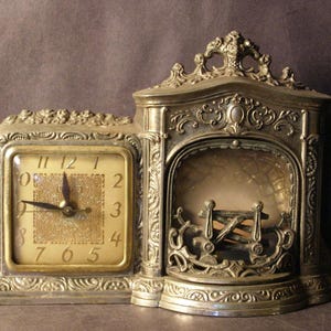 May include: An antique silver-toned clock with an ornate design. The clock face is square with gold numerals and hands. The clock is attached to a decorative fireplace-shaped structure.