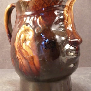 May include: A brown ceramic pitcher with a face design. The pitcher has a handle and a spout. The face is detailed with a nose, eyes, and mouth. The colors are shades of brown and tan.
