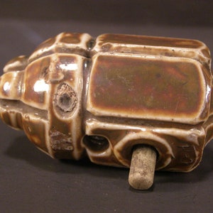 May include: A brown ceramic insulator with a wooden peg. The insulator has a textured surface and is shaped like a rounded rectangle.