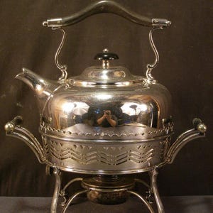 May include: A silver-colored tea kettle with a black handle and lid knob, set on a matching stand. The stand has decorative cutouts and curved legs. The kettle has a spout and a curved handle.
