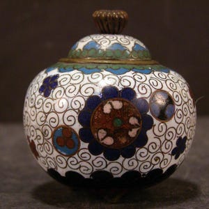 Puede incluir: Un pequeño tarro redondo de cerámica blanca con un patrón floral azul, verde y rojo. El tarro tiene una tapa con un pomo decorativo.