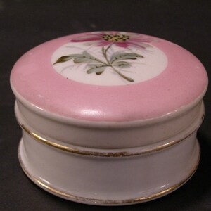 May include: A small, round, white porcelain trinket box with a pink lid. The lid is decorated with a hand-painted floral design in shades of pink, green, and white.