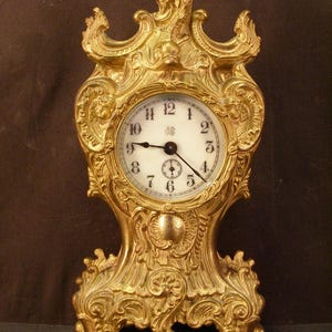 May include: Ornate gold-colored mantel clock with a white clock face and black hands. The clock features elaborate scrollwork and decorative details. The clock's face has black Roman numerals and a small seconds dial. The clock is a vintage style.