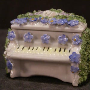 May include: A white porcelain piano trinket box decorated with blue and yellow flowers. The box is shaped like a piano with a lid that opens to reveal a small interior space.