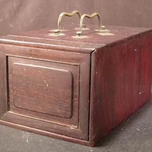 May include: A rectangular wooden box with a dark reddish-brown finish. It features a recessed panel on the front and two brass handles on the top. The box appears to be vintage or antique.