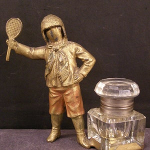 May include: A gold-toned figurine of a person holding a tennis racket, standing next to a glass inkwell with a gold-toned base.