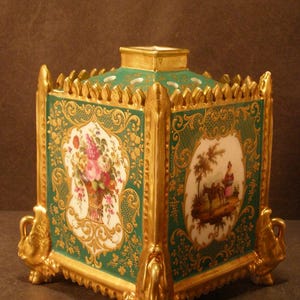May include: A square porcelain potpourri jar featuring a deep green background with intricate gold filigree detailing. The four sides display hand-painted scenes of floral bouquets and figures. It has a gold-trimmed top and four ornate feet.