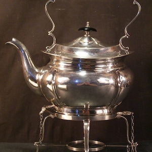 May include: A silver-plated tea kettle with a black handle and lid finial. The kettle sits on a matching stand with decorative legs and chains. The spout curves gracefully, and the overall design is ornate and elegant.