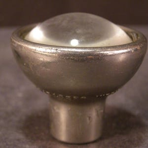 May include: A vintage, silver-toned cabinet knob with a clear, rounded glass top. The knob has a bowl-shaped body and a cylindrical base. The metal shows signs of wear, suggesting age and use. The knob is designed for furniture.