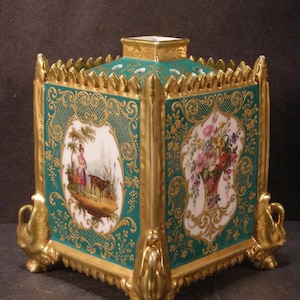 May include: A square porcelain potpourri jar featuring a teal green body with ornate gold filigree and gilded edges. The sides display hand-painted oval vignettes of a person with an animal and a floral bouquet. It has four gold swan-shaped feet.