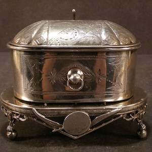 May include: A silver-toned metal box with an ornate, engraved lid and a central ring pull handle on the front. The box rests on a decorative oval stand featuring four small feet and a central circular emblem flanked by two crossed sword-like motifs.
