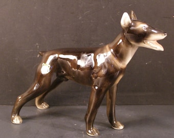 May include: A ceramic figurine of a dog in a standing pose. The dog has a glossy, dark brown coat with lighter brown and white accents on the ears, snout, and paws. The dog's mouth is open, and it appears to be barking.