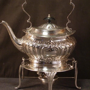 May include: Ornate silver tea kettle with a black handle and lid finial. The kettle sits on a matching stand with decorative legs and a small burner. The design features detailed scrollwork and a curved spout.